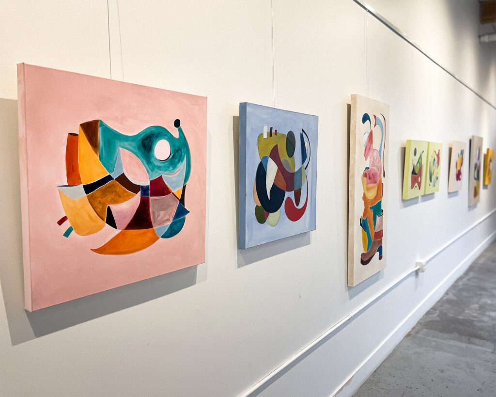 A collection of abstract paintings in various sizes and colors displayed on a white wall in a gallery setting.