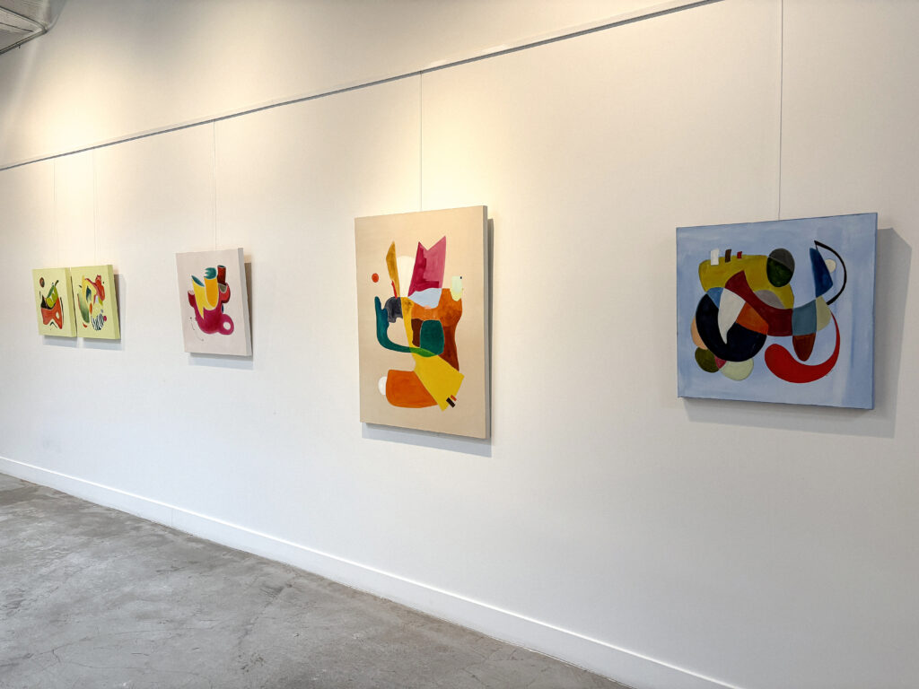 Four abstract paintings hang on a white wall in a gallery. The paintings feature bold shapes and colors against light-colored backgrounds.