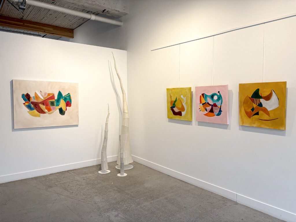 A gallery wall displays abstract paintings and sculptures. On the left, a large, light-colored canvas features a colorful abstract design. To its right, three tall, white, textured sculptures stand on the floor. Further to the right, three smaller square canvases with vibrant abstract shapes are hung in a row on a white wall.