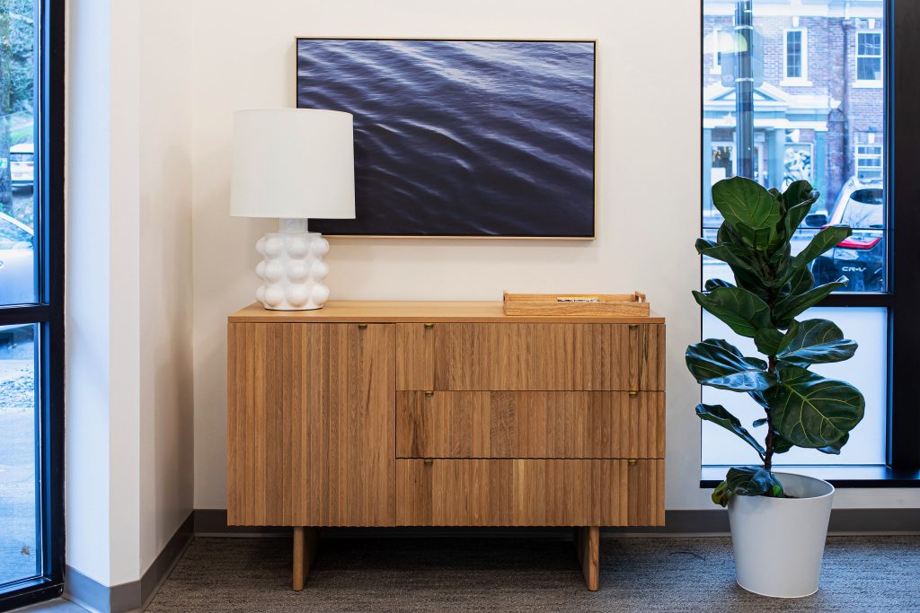 A modern wooden credenza with a white bubble lamp and abstract art of water waves above it, with a potted fiddle leaf fig plant to the right.