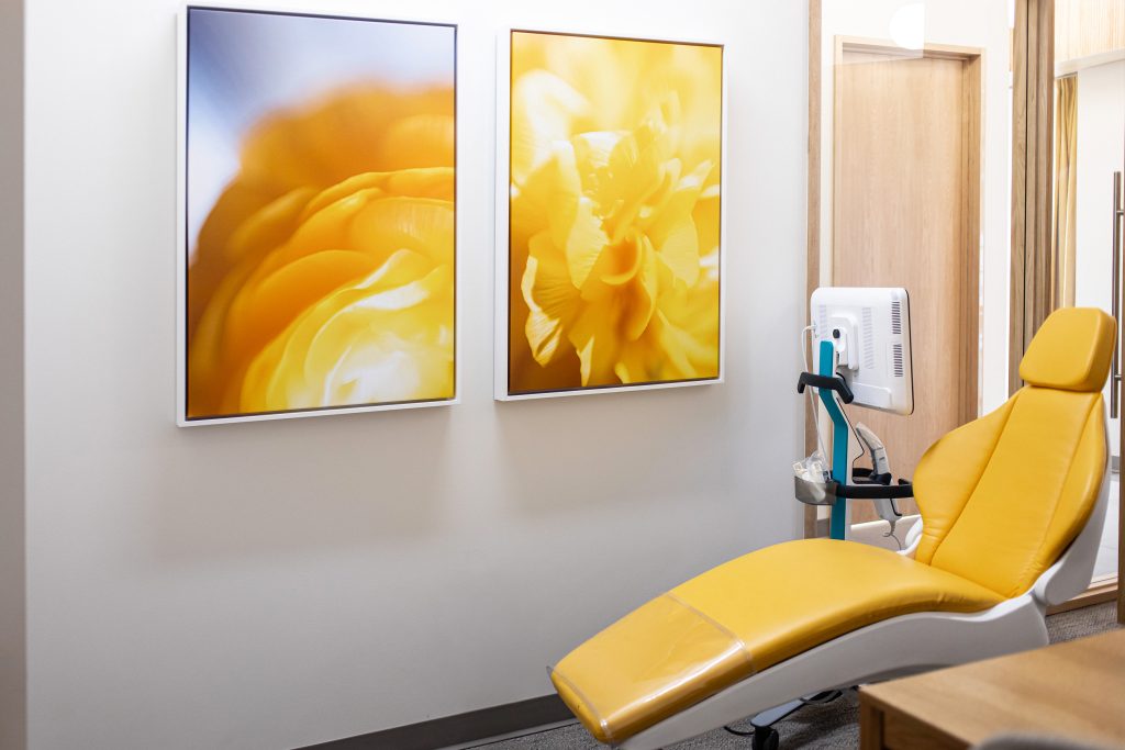 Two framed close-up images of yellow flowers hang on a wall above a bright yellow dental chair. A medical device is attached to a pole next to the chair.