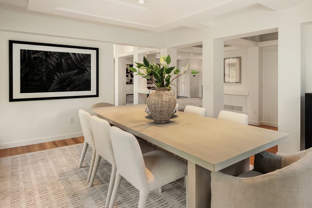 A dining room with a long, light wood table set with white chairs and a vase of greenery in the center. A large black and white framed artwork hangs on the wall above a patterned rug.